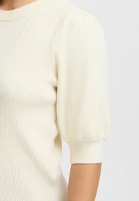 Kremkrāsas ribota adīta džemperis ar īsām pūkainām piedurknēm un noapaļotu kakla izgriezumu. Virsma ir maiga un nedaudz elastīga.