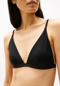 Crni trokutasti bralette s tankim naramenicama i elastičnom trakom ukrašenom logom. Glatka tkanina s mat završetkom. Minimalistički dizajn.