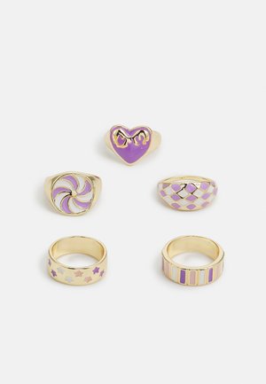 Cinco anéis em metal de tom dourado. As características incluem um design de coração roxo, um padrão em espiral, motivos de diamantes e estrelas, em cores pastel.