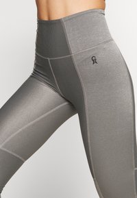 Legginsy w kolorze szarym, o wysokim stanie, wykonane z teksturowanej, elastycznej tkaniny z delikatnym połyskiem. Posiadają detale szwów oraz małe logo w pobliżu pasa.