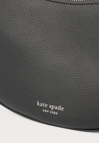 Fekete bőr kézitáska texturált felülettel, ívelt formával és alul fehér "kate spade NEW YORK" logóval.