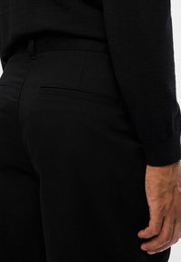 Close-up van een persoon in zwarte broek en een zwart langstpulptop, met zicht op de heup, achterzak en de rechterhand ontspannen langs het lichaam.