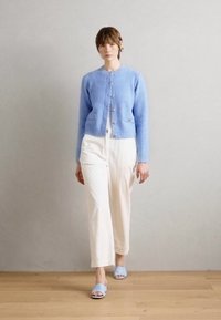 Helle blaue Strickjacke mit rundem Halsauschnitt und Knöpfen, kombiniert mit weißen weit geschnittenen Hosen und hellblauen Sandalen an einem Modell.