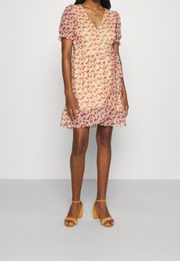 Blommig klänning med V-ringning, puffärmar och insnörning vid midjan i orange, rött och krämfärgade toner. Matchas med gula klacksandaler.