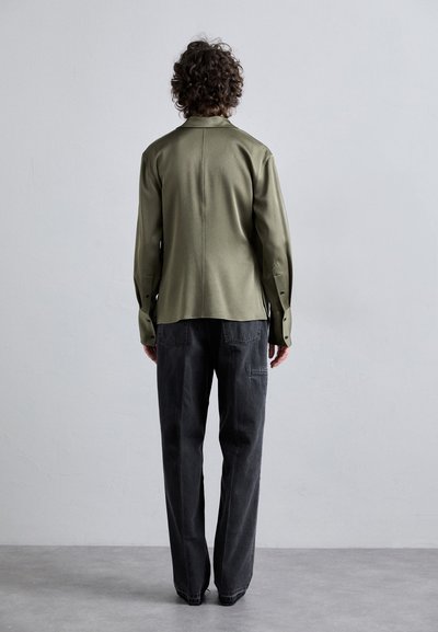 Olivegrünes Hemd mit geradem Schnitt und seitlichen Schlitzen, kombiniert mit schwarzen Jeans mit Taschen und geradem Bein.