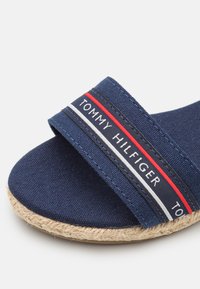 Marineblauwe stoffen slidesandaal met een geweven jute zool en een gestreepte elastieken band met "TOMMY HILFIGER" in witte, rode en marineblauwe accenten.