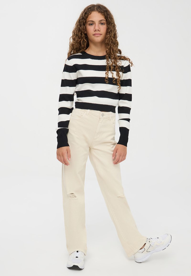 Maglione a righe bianco e nero a costine, pantaloni beige a gamba larga con dettagli strappati e sneakers bianche con accenti neri.