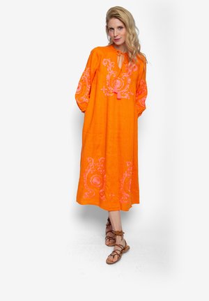 Kobieta stojąca ze skrzyżowanymi nogami, ubrana w pomarańczową sukienkę midi z różowym haftem, z dekoltem wiązanym frędzlami, i brązowe sandały na sznurowadła.