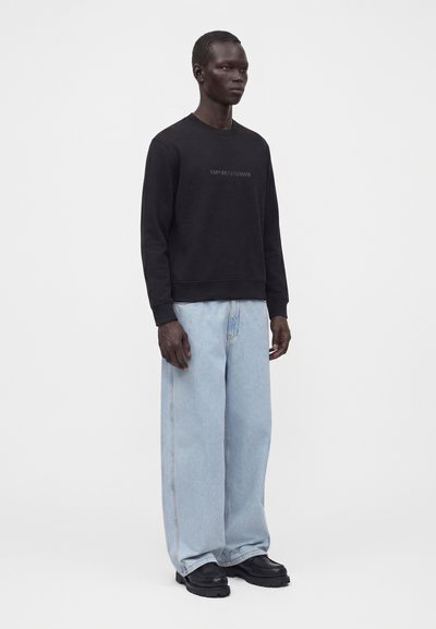 Homme debout portant une chemise noire à manches longues avec logo, un jean bleu clair à jambes larges et des chaussures noires sur un fond uni clair.