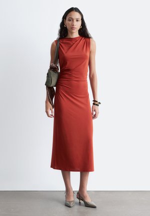 Vrouw draagt een mouwloze midi-jurk in roestkleur, grijze kitten heels, met een groene handtas met een polkadot sjaal, staand tegen een witte muur.