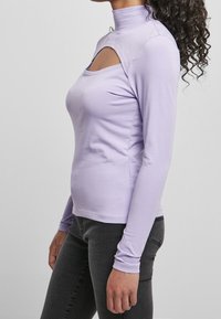 Světle fialová košile s dlouhým rukávem a vysokým límcem, s výřezem na hrudi. Měkká látka s přiléhavým střihem, doplněná tmavými džínami.