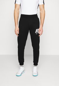 Zwarte joggingbroek met elastische taille en boorden, met een wit logo aan de zijkant. Combineert met witte sportschoenen en grijze sokken.