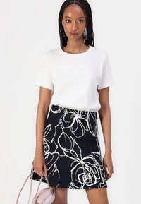 Schwarzer Rock mit weißem Blumenmuster, kombiniert mit einem weißen T-Shirt mit Rundhalsausschnitt. Das Model hält eine hellrosa Tasche. Glatte Textur, taillierte Passform.