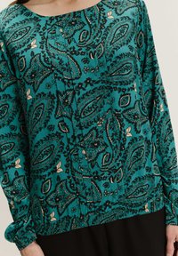 Blusa verde azulado con dobladillo y puños fruncidos, que presenta un intrincado patrón en negro y beige de paisley y flores en la tela.
