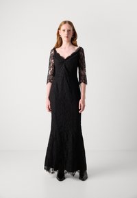Robe en dentelle noire avec des manches longues, décolleté en V et corsage ajusté. Présente une partie inférieure évasée avec un ourlet festonné et des motifs en dentelle élaborés.