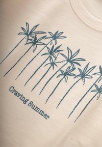 Camiseta de algodón de color crema, con un diseño de palmera en color azul verdoso y el texto "Deseando Verano" en una fuente sencilla debajo del gráfico.