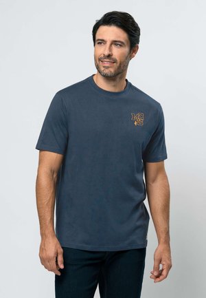 Sötét hajú és szakállas férfi, tengerészkék pólót visel, amelyen narancssárga "MCS" logó látható a mellkasán, egyszínű világos háttér előtt áll.