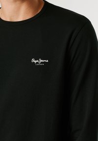 Črna majica z dolgimi rokavi in majhnim belim napisom "Pepe Jeans London" na levi strani prsnega koša, ki jo nosi oseba.