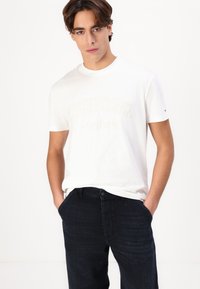 Hvit bomull T-skjorte med "TOMMY JEANS New York"-logo i hevet kremfarge, kombinert med mørkeblå denim jeans. Enkel, uformell design.