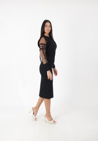 Zwarte aansluitende jurk met lange, doorzichtige mesh-mouwen met ronde uitsparingen. Model draagt open teen, gehakte muiltjes met een geruit patroon.