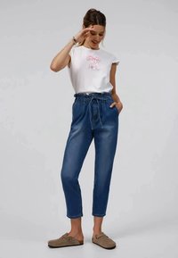 Biely top s krátkym rukávom a ružovým mašľovým akcentom, kombinovaný s modrými džínami s vysokým pásom. Model má na sebe béžové šľapky.