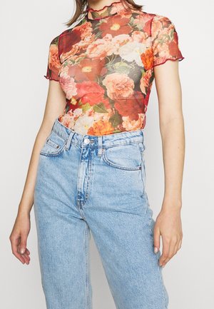 Frau trägt ein durchsichtiges, kurzärmeliges Oberteil mit roten, orangefarbenen und pinken Blumen, das in eine hoch taillierte, hellblaue Jeans aus Denim gesteckt ist.