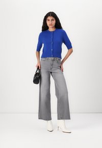 Cardigan corto azul de manga corta con cierre de botones, combinado con unos pantalones de mezclilla gris de pierna ancha de cintura alta y botas blancas hasta el tobillo. Sosteniendo un bolso negro.