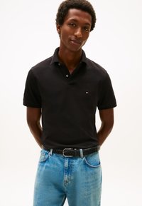 Polo nera con maniche corte e colletto, con un piccolo logo del marchio. Indossata con jeans di denim azzurro chiaro e una cintura nera intrecciata.