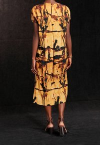 Vestido de estampado floral amarillo con mangas cortas, un corte holgado y largo hasta la rodilla. Incluye una abertura en la espalda y contrasta con un fondo oscuro.