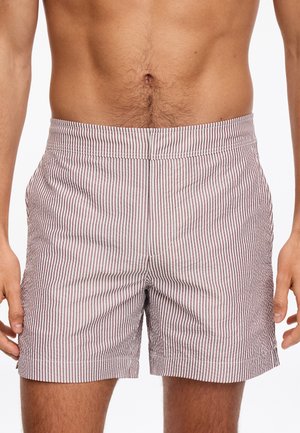 Mężczyzna noszący czerwone i białe kąpielówki w pionowe paski, pokazany od talii do połowy uda na jednolitym tle.