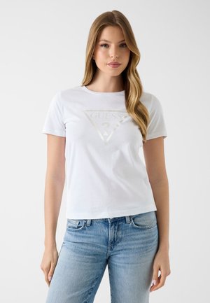 Jeune femme aux longs cheveux blonds portant un t-shirt blanc avec le logo Guess et un jean bleu clair, debout devant un fond blanc uni.