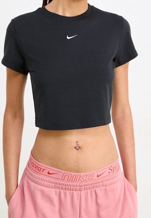 Kobieta ubrana w czarny krótki t-shirt Nike i różowe dresy Nike z logo na pasie, widoczny piercing w pępku.