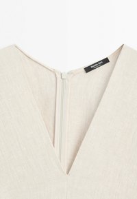 Zbliżenie na dekolt sukienki z beżowej, teksturowanej tkaniny z centralnym zamkiem błyskawicznym z tyłu i widoczną metką marki Massimo Dutti.