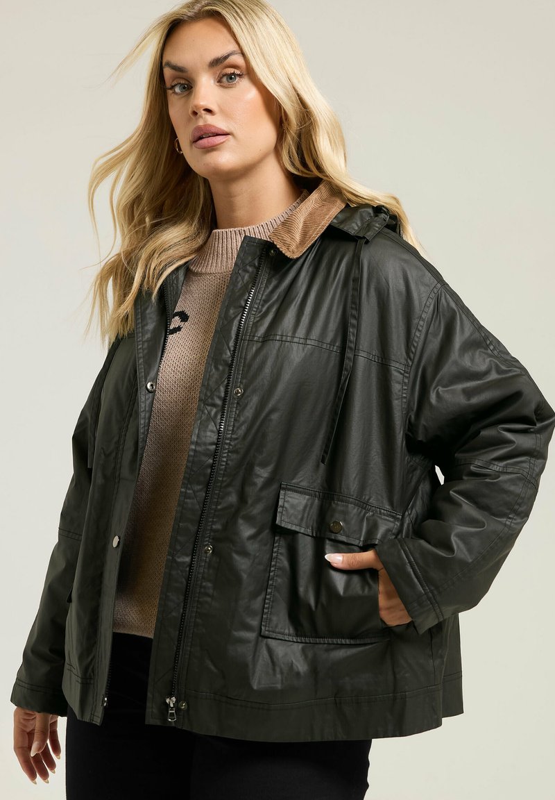 Veste imperméable vert foncé avec une fermeture éclair à l'avant, des poches à boutons et un col en velours côtelé marron. Les cordons de la capuche sont visibles, montrant un ajustement décontracté.