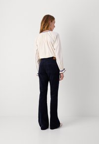 Weiße kurz geschnittene Bluse mit blauer Stickerei am Ausschnitt, kombiniert mit dunkelblauen ausgestellten Cordhosen. Glatte Textur, tailliertes Design.