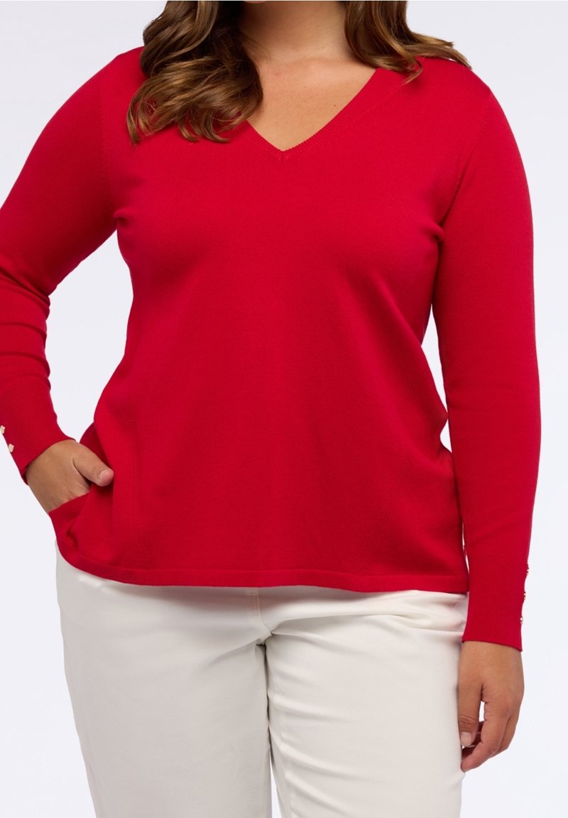 Femme portant un pull rouge à manches longues col V avec des boutons sur les manches, associé à un pantalon blanc, debout avec une main dans la poche.