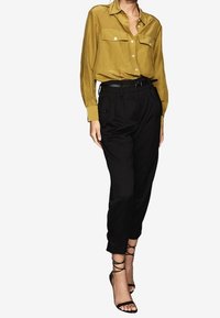 Blouse en soie couleur moutarde avec deux poches avant et une ceinture noire, associée à un pantalon noir et des talons noirs à lanières.