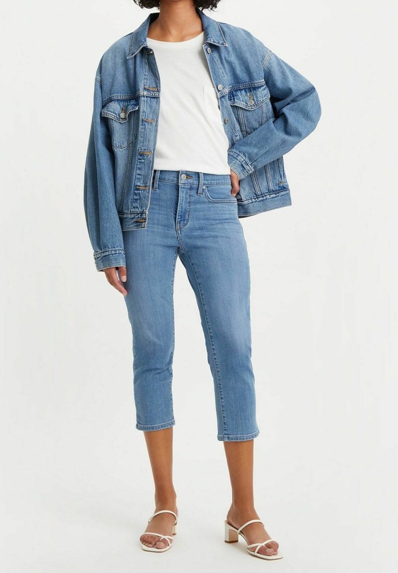 Chaqueta de mezcla vaquera azul claro sobre una camiseta blanca, combinada con unos pantalones vaqueros cortos azul claro y sandalias de tacón blancas. La chaqueta tiene dos bolsillos en el pecho.