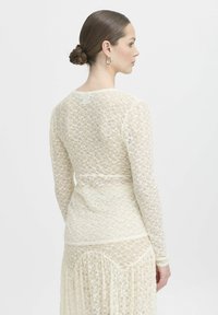 Top in pizzo bianco sporco con maniche lunghe, silhouette aderente e delicato motivo floreale. Presenta orlo e scollatura ondulati.