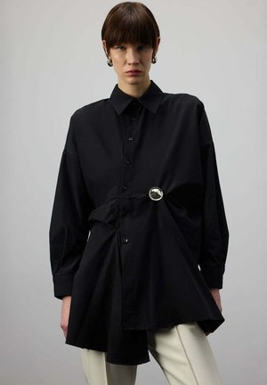 Άτομο που φοράει υπερμεγέθη μαύρο πουκάμισο με κουμπιά, σφιγμένο στη μέση με ασημένια κυκλική αγκράφα, σε συνδυασμό με ανοιχτόχρωμο παντελόνι.