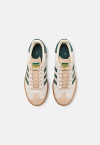 Les baskets Adidas présentent une tige beige avec des rayures vertes et jaunes, des accents en daim et une semelle en caoutchouc. Les lacets sont beiges avec un col rembourré.