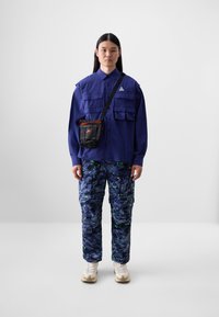 Mørkeblå knappeskjorte med brystlommer, kamuflasje cargo-bukser i blått med grønne detaljer, og en liten svart crossbody-veske med oransje kant.