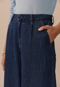 Donkerblauwe denim hoge taillebroeken met voorplooien, zijzakken en contrasterende beige stiksels. Glad textuur, relaxte pasvorm.