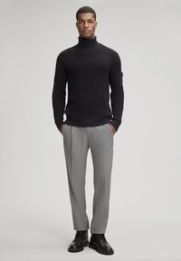 Pull en maille côtelée noir à col roulé, assorti à un pantalon plissé gris clair et des bottes noires à lacets. Textures lisses et détails de coupe ajustée.