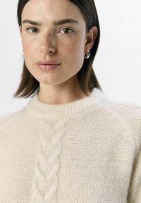 Creme kabelstrikket sweater med rund hals og ribbede manchetter. Blød tekstur og afslappet pasform med en central fletdetalje.