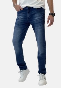 Vaqueros de mezclilla azul oscuro con un ajuste Slim, que presentan un leve desgaste y detalles de costura. Combinados con zapatillas blancas para un look informal.