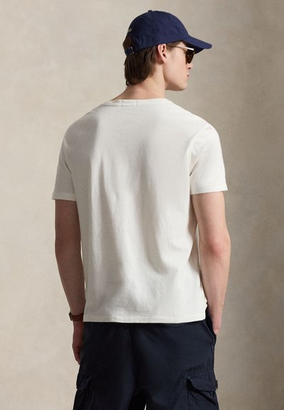 Man die een wit T-shirt met korte mouwen, marineblauwe cargoshorts, een marineblauwe baseballpet en een zonnebril draagt, kijkend met zijn rug naar een effen achtergrond.