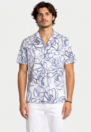 Uomo che indossa una camicia bianca a maniche corte con bottoni e motivi floreali blu a linee e pantaloni bianchi, in piedi su uno sfondo chiaro uniforme.