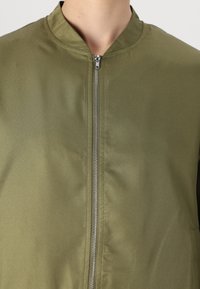 Giubbotto bomber verde oliva con chiusura a zip, colletto rotondo e tessuto liscio, leggermente lucido. Design semplice e moderno.