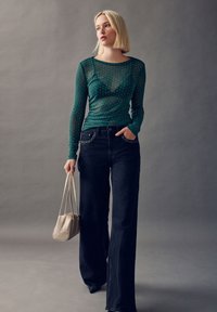 Grøn netlangærmet top med en tætsiddende pasform, pyntet med små prikker, kombineret med højtaljede mørke jeans og en beige håndtaske.